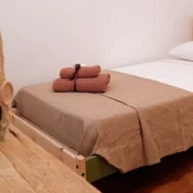Habitación minimalista con cama individual, sábanas blancas y una manta beige. En la cama hay toallas enrolladas en tonos terracota. Un frasco decorativo con piedras y elementos naturales se encuentra sobre una mesa de madera.