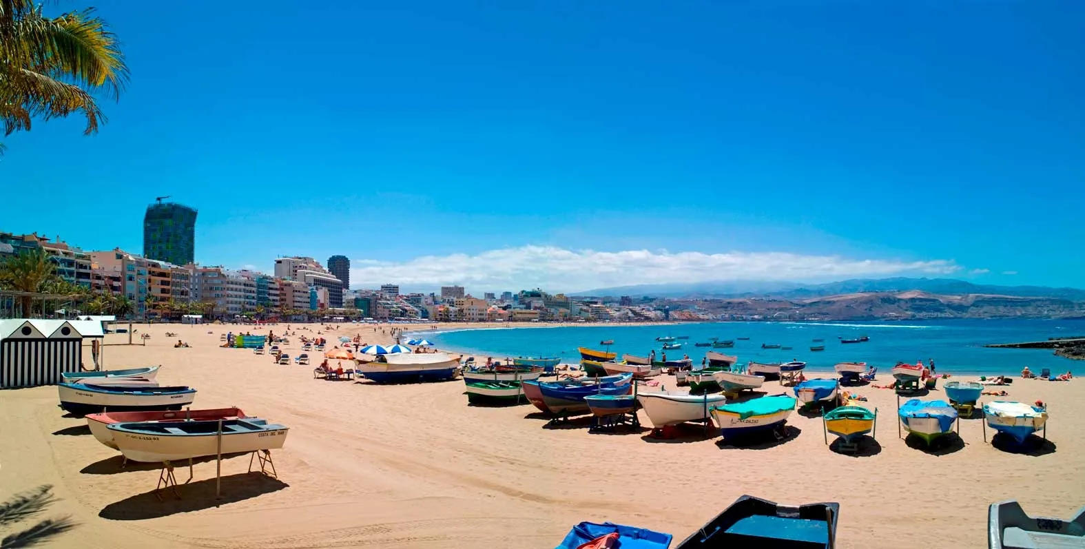Playa soleada con embarcaciones en la arena, edificios de fondo y personas disfrutando del día. Ideal para turismo y actividades acuáticas.