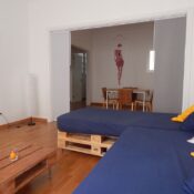 Sala de estar moderna con sofá de pallets y cojines azules, mesa de comedor de madera en el fondo, decoración minimalista y luminosa.