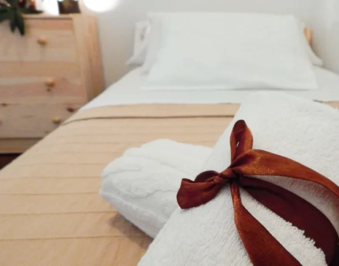 Toalla blanca decorada con un lazo marrón, sobre una cama con sábanas beige y un cabecero sencillo. Al fondo, se puede ver un mueble de madera y elementos decorativos que aportan calidez al ambiente.