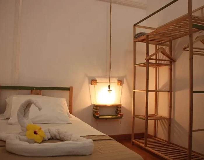 Habitación acogedora con cama sencilla, decoración de toalla en forma de cisne y flor amarilla. Se observa un armario de madera y una lámpara colgante, creando un ambiente cálido y minimalista. Ideal para estancias cómodas y relajantes.