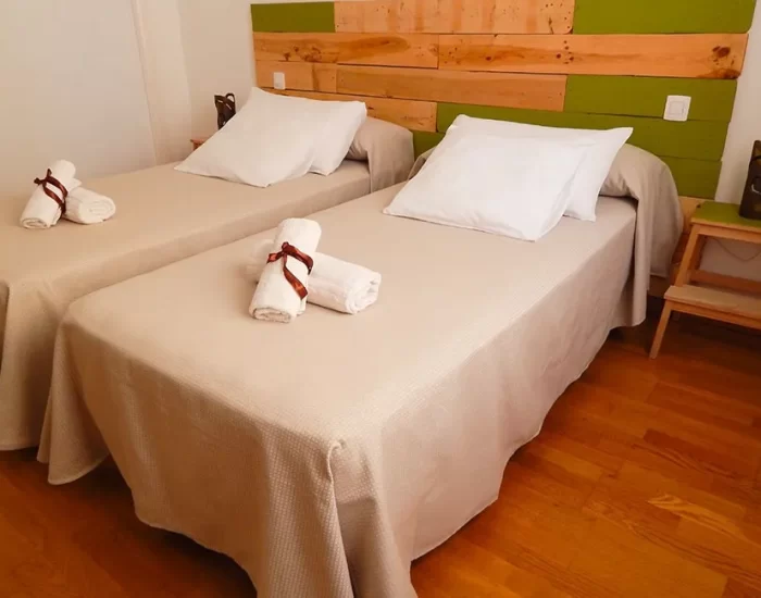 Habitación acogedora con dos camas individuales, decorada con un cabecero de madera y tonos verdes. Incluye toallas enrolladas y almohadas blancas, ideal para una estancia relajante. Suelo de madera que aporta calidez al ambiente.
